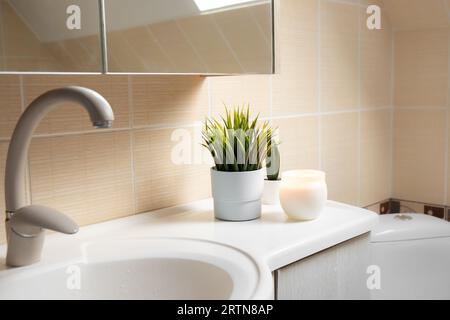 Uso di fiori artificiali nell'interno del bagno a casa. Pianta di fiori in vaso nell'angolo del lavandino del bagno con candele a fuoco. Foto Stock