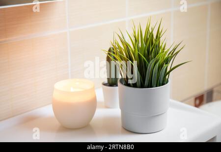 Uso di fiori artificiali nell'interno del bagno a casa. Pianta di fiori in vaso nell'angolo del lavandino del bagno con candele a fuoco. Foto Stock