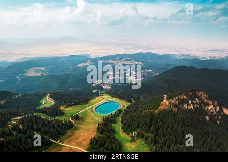 Veduta aerea di Poiana Brasov, Romania. Lago situato in cima alla montagna coperta da una lussureggiante foresta, città in lontananza Foto Stock