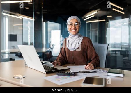 Ritratto di una giovane donna araba di successo in hijab che lavora in ufficio con documenti e laptop. Sorridere e guardare la telecamera. Foto Stock