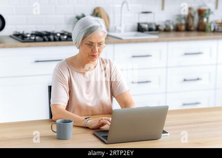 Donna caucasica matura con capelli grigi focalizzati e bicchieri, sedersi a una scrivania a casa in cucina con un computer portatile, lavorare in remoto, inviare messaggi con un cliente, lavorare su un progetto, guarda concentrato sullo schermo Foto Stock