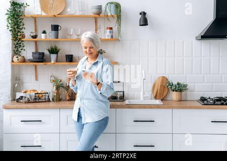 Gorgeous felice caucasico medio vecchio grigio capelli donna, stand a casa in cucina, tiene smartphone e un bicchiere di caffè, messaggi nei social network, la lettura di notizie, fa un ordine online, sorrisi Foto Stock