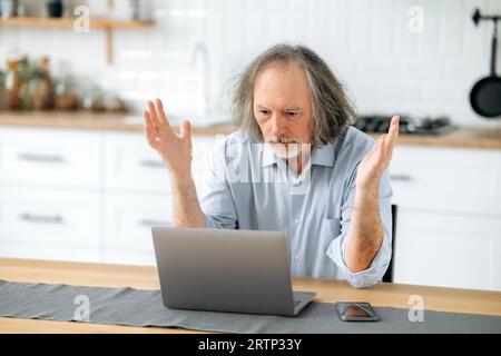 Frustrato uomo anziano dai capelli grigi caucasici, pensionato, mentore finanziario, siede a un tavolo in cucina, guarda confusamente a un computer portatile, è perplesso dalla notizia o dal risultato del lavoro, gesturando le mani Foto Stock