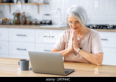 Soddisfatto donna caucasica di mezza età con capelli grigi con occhiali, seduto a casa in cucina al tavolo, guarda lo schermo del computer portatile, vuole qualcosa, si preoccupa, tiene le palme insieme, prega Foto Stock