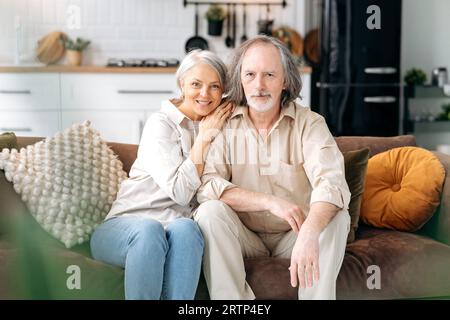 Felice insieme. Pensionati moderni. Coppia di sposi caucasici di mezza età, sposi dai capelli grigi trascorrono del tempo insieme nel soggiorno, moglie e marito si siedono su un divano, abbracciando, guardando la macchina fotografica, sorridendo Foto Stock