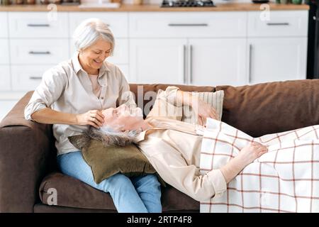 Due felici pensionati caucasici, gioiosi sposi e sposi maturi, trascorrono del tempo insieme a casa sul divano del soggiorno, l'uomo giace sulle gambe della donna, si parlano e sorridono Foto Stock