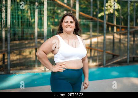 Ritratto di una donna sorridente in sovrappeso con una tuta da ginnastica su un campo sportivo in un parco che guarda nella macchina fotografica Foto Stock