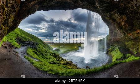 Maestosa grotta naturale con cascata Seljalandsfoss che scorre e un campo lussureggiante durante l'estate in una giornata cupa nel sud dell'Islanda Foto Stock