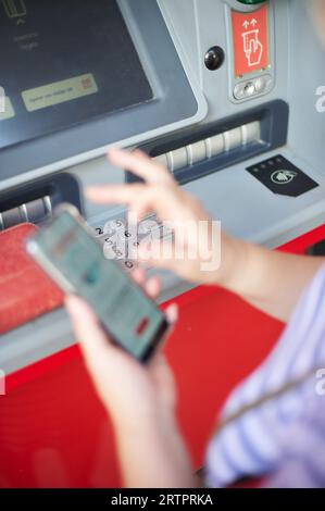 Donna irriconoscibile che tiene un telefono cellulare davanti a un bancomat bancario che svolge un'operazione di online banking. Foto Stock