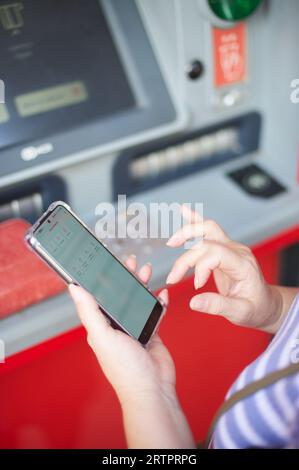 Donna irriconoscibile che tiene un telefono cellulare davanti a un bancomat bancario che svolge un'operazione di online banking. Foto Stock