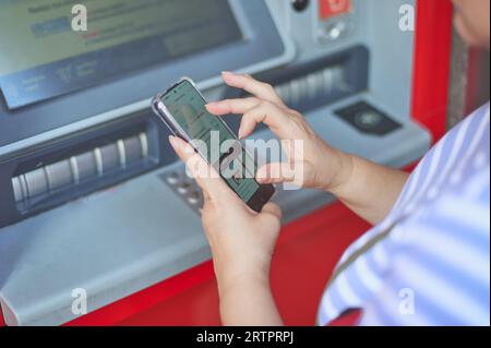 Donna irriconoscibile che tiene un telefono cellulare davanti a un bancomat bancario che svolge un'operazione di online banking. Foto Stock