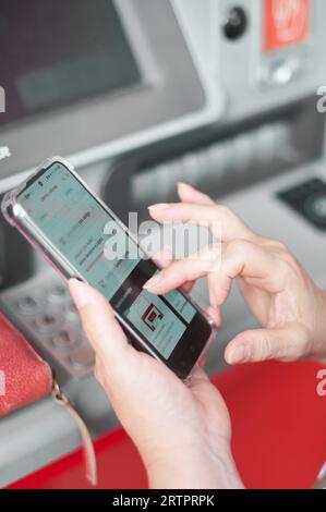 Donna irriconoscibile che tiene un telefono cellulare davanti a un bancomat bancario che svolge un'operazione di online banking. Foto Stock