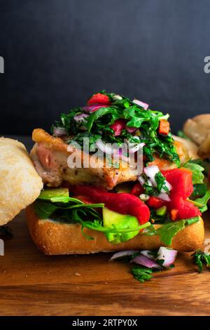 Sandwich di pollo Chimichurri su tagliere di legno: Panino aperto con avocado, peperoni arrostiti e rucola Foto Stock