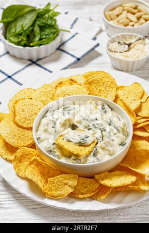 salsa cremosa di carciofi spinaci alle mandorle con patatine fritte su piatto bianco su tavola di legno bianco con ingredienti, vista verticale dall'alto, primo piano Foto Stock