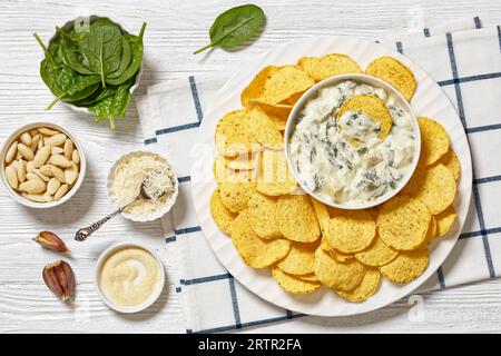 salsa cremosa di carciofi spinaci alle mandorle con patatine fritte su piatto bianco su tavola di legno bianco con ingredienti, vista orizzontale dall'alto, posa piatta Foto Stock