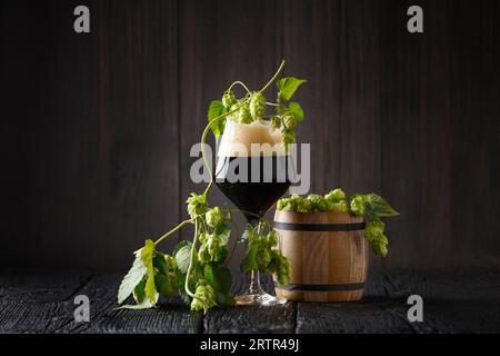 Tazza di vetro di birra scura, rami di luppolo e botte di legno con luppolo su sfondo di legno scuro. Tradizione della produzione di birra. Oktoberfest. Festival della birra. St Foto Stock