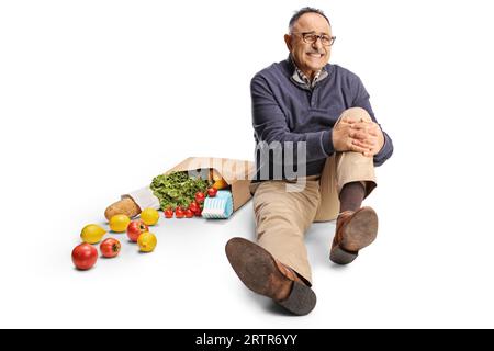 Uomo maturo con alimentari che tiene il ginocchio doloroso e seduto a terra isolato su sfondo bianco Foto Stock