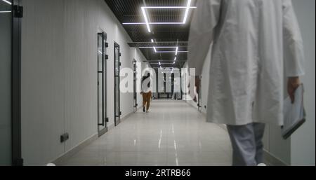 Medico maturo, operatore sanitario in camice bianco da laboratorio e con cartelle di carte cammina lungo il luminoso corridoio della clinica. Personale medico e persone in ospedale o centro medico. Vista posteriore. Foto Stock