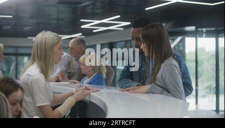 Coppie multietniche si trovano vicino alla reception nell'area della hall della clinica e prendono appuntamento con un medico. L'amministratore donna parla con i pazienti al banco informazioni. Il personale medico lavora in background. Foto Stock