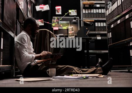 Agente investigativo privato che analizza le foto di sorveglianza sul portatile. Detective afroamericano seduto sul pavimento e lavorando fino a tardi studiando le fotografie della scena del crimine al computer Foto Stock