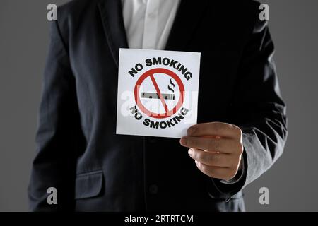 Uomo che tiene una carta con un cartello di divieto di fumo su sfondo grigio, primo piano Foto Stock