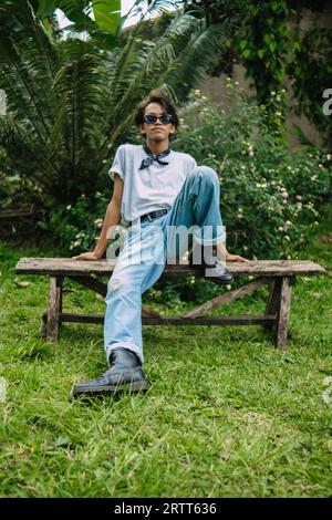 Un uomo asiatico con occhiali da sole e una maglietta bianca siede su un tavolo di legno in un parco la mattina Foto Stock