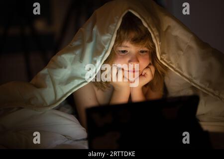 Bambino che guarda il tablet prima di dormire. Il bambino legge l'e-book. Il ragazzino guarda lo schermo del tablet in alto. Ragazzo che gioca un tablet sdraiato su un Foto Stock