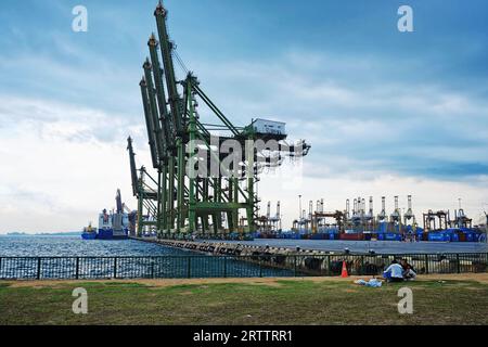 Due pescatori ricreativi che si accovacciano sulla spiaggia davanti a una fila di gru a cavalletto verdi al Tanjong Pagar Terminal Singapore Foto Stock