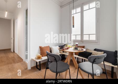 Piccolo tavolo da pranzo in legno con colazione per due e vino con due sedie e una panca con cuscini nell'angolo contro una parete bianca e un grande Foto Stock