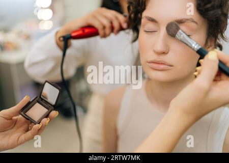 Primo piano ritagliato di una truccatrice femminile irriconoscibile che applica l'evidenziatore per la scultura del viso di una bella giovane donna in uno studio di bellezza moderno. Foto Stock