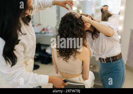 Vista posteriore di una truccatrice che si prepara ad applicare il trucco a una giovane donna seduta mentre una parrucchiera femminile si prepara ad acconciare in un salone di bellezza, primo piano, posteriore Foto Stock