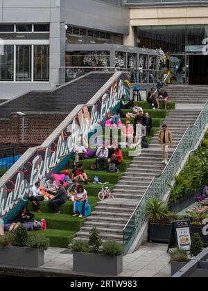 Persone che si divertono a mangiare all'aperto su alcuni gradini ricoperti di erba presso l'edificio Mailbox di Birmingham, Regno Unito Foto Stock