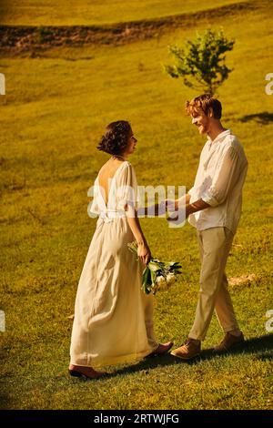 sposo di rosse e sposa asiatica con fiori che indossano un elegante abbigliamento boho e si tengono per mano in campo Foto Stock