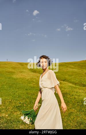 incantevole sposa asiatica in stile boho che cammina con bouquet nuziale su prato verde, ambiente rurale Foto Stock