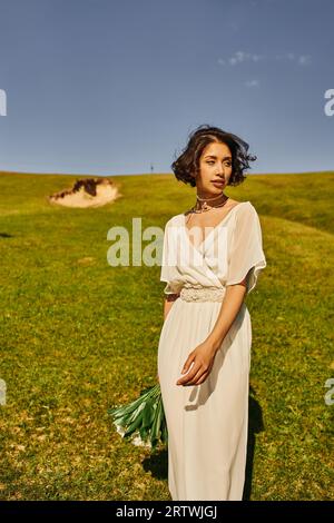 bruna donna asiatica con abito da sposa in stile boho che regge fiori e guarda lontano nel verde Foto Stock