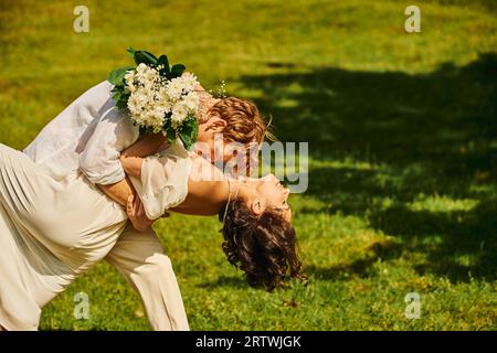 giovane sposo rosso che abbraccia e bacia la sposa asiatica con fiori su prato verde, matrimonio rustico Foto Stock