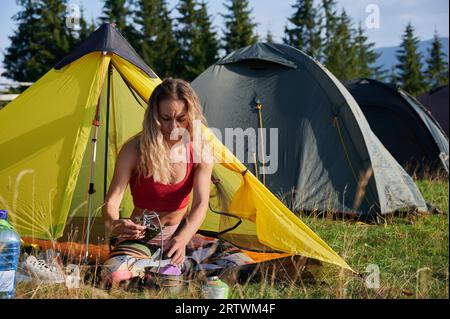 Campeggiatore all'aperto. Escursioni turistiche di sesso femminile in estate. Bella donna che indossa abbigliamento sportivo, seduta sull'erba, che cucina, mette pentole. Concetto di turismo e avventura. Foto Stock