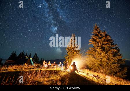 Campeggio notturno in montagna sotto il cielo stellato. Gruppo di persone che viaggiano, camminano sulle colline. Amici seduti sull'erba in campeggio, a parlare, ad ammirare il fuoco. Concetto di turismo, escursionismo e avventura. Foto Stock