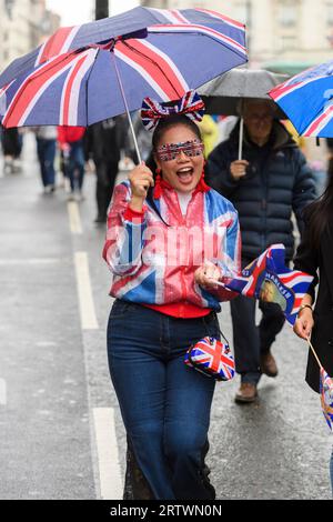 Una donna, vestita con una giacca Union Jack, e occhiali, e con un ombrello e una borsa coordinati, cammina lungo un Piccadilly piovoso, parte di una grande folla Foto Stock