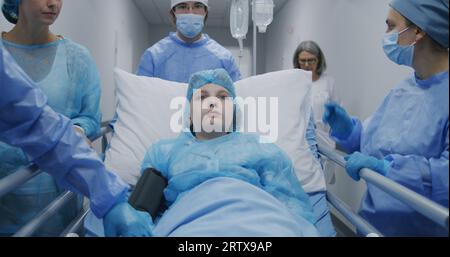 Medici e paramedici spingono la barella con un bambino malato in sala operatoria e parlano con lei. Il medico controlla il pulsossimetro sulla mano del paziente. Il personale medico lavora in clinica. Vista posteriore. Ripresa di tracciamento. Foto Stock