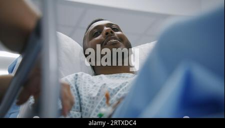 L'uomo afro-americano giace sulla barella e parla con i medici prima dell'operazione. Il medico si trova vicino alla barella con il paziente nel centro medico. Il personale medico lavora nel pronto soccorso. Primo piano. Foto Stock