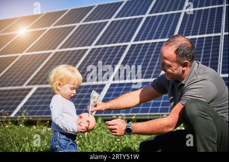 Padre e suo figlio mettono soldi in salvadanaio per risparmiare sui pannelli solari. Ragazzino interessato a risparmiare denaro per il futuro. Concetto di investimento nelle energie rinnovabili. Foto Stock