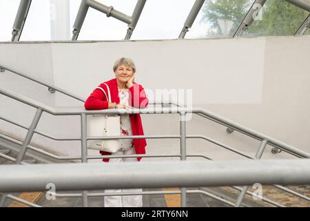 Donna matura di 70 anni in abiti casual in posa in Underground Crossing. Sorridente bella vera femmina con i capelli grigi regge la ringhiera. Pensione Foto Stock