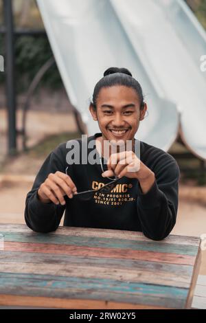 Un uomo asiatico con i capelli legati e indossando una giacca maglione nera è seduto a rilassarsi al tavolo di un caffè quando incontra i suoi amici nel pomeriggio Foto Stock