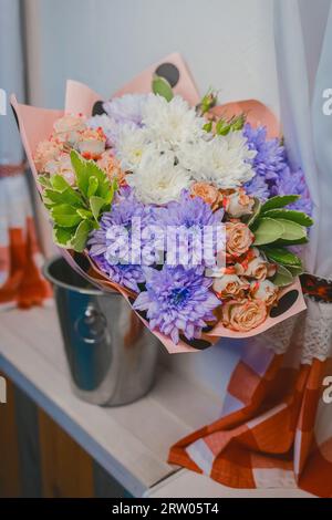 Un bouquet di fiori, crisantemi viola e bianchi e rose color crema si trovano in un secchio di metallo nell'acqua sullo sfondo dell'inter Foto Stock