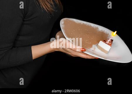 Donna in abiti scuri con manicure rossa che tiene un piatto bianco di congratulazioni con cheesecake e una candela di compleanno in fiamme. Foto Stock