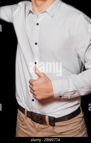 La mano di un ragazzo con una camicia bianca e pantaloni beige mostra la classe con i pollici in alto su uno sfondo scuro. Foto Stock