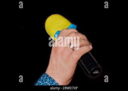 Il primo piano della mano di un uomo contiene un microfono con un bordo giallo morbido, uno strumento di canto karaoke su sfondo scuro. Foto Stock