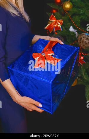 Una ragazza tiene una scatola in un film regalo blu regalo di Capodanno sullo sfondo di un albero di abete rosso. Foto Stock