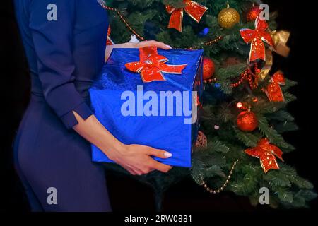 Una ragazza tiene una scatola in un film regalo blu regalo di Capodanno sullo sfondo di un abete rosso, primo piano. Foto Stock